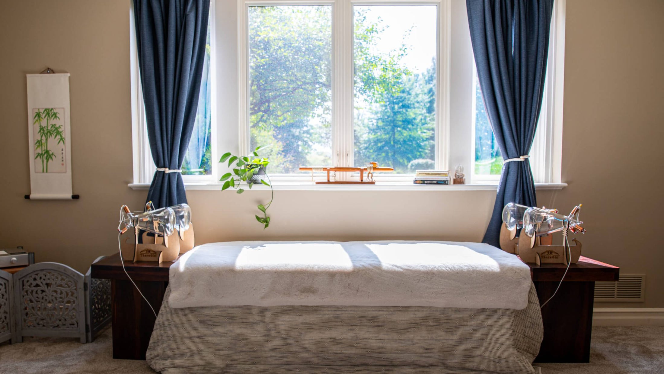 Bedroom with a bed, theraphi bulbs on nightstands, and large windows with blue curtains.